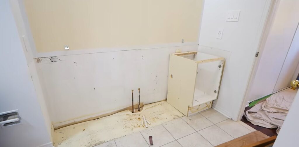 An empty bathroom wall showing exposed plumbing and a single partially installed cream-colored cabinet on a tiled floor.