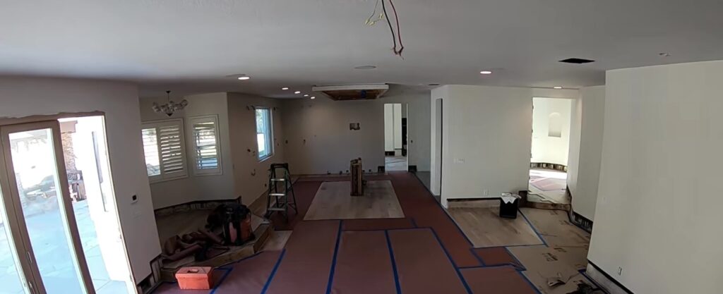 A wide-angle view of a large open-concept living and kitchen area during renovation, featuring protective brown paper taped to the floors and a central area with newly installed light wood flooring.