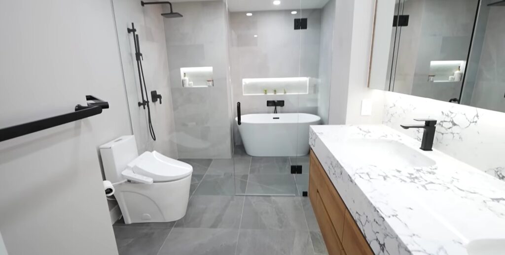 A finished contemporary bathroom featuring gray floor tiles, a white freestanding soaking tub inside a glass shower enclosure, a white bidet toilet, and a marble-topped vanity with black hardware.